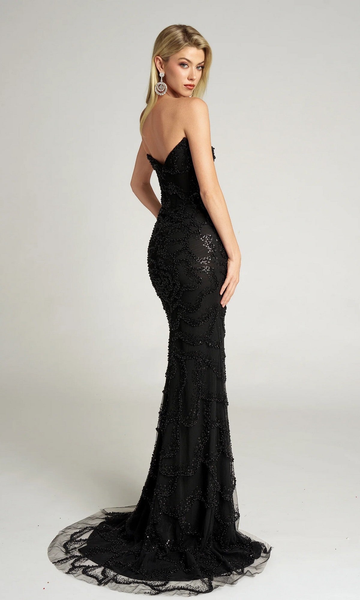 A woman in the Portia and Scarlett PS26126 long formal dress, featuring beaded details and a small train, stands facing away and looks over her shoulder against a plain background.
